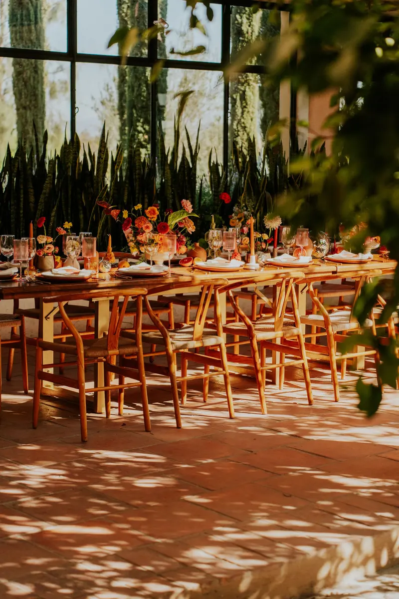 Mesa larga con flores y velas en una galería con plantas, bajo luz cálida